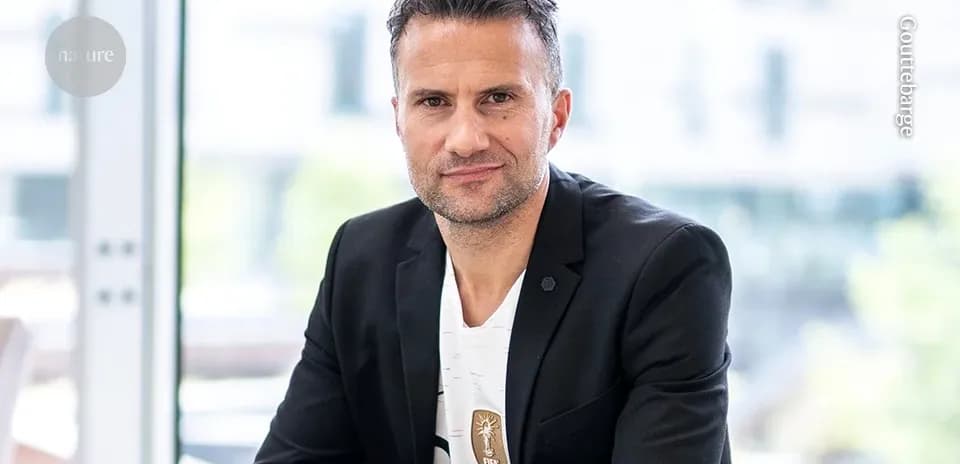 Man in black blazer sitting indoors, looking at camera