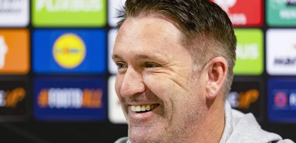 Coach smiling during press conference with logos behind